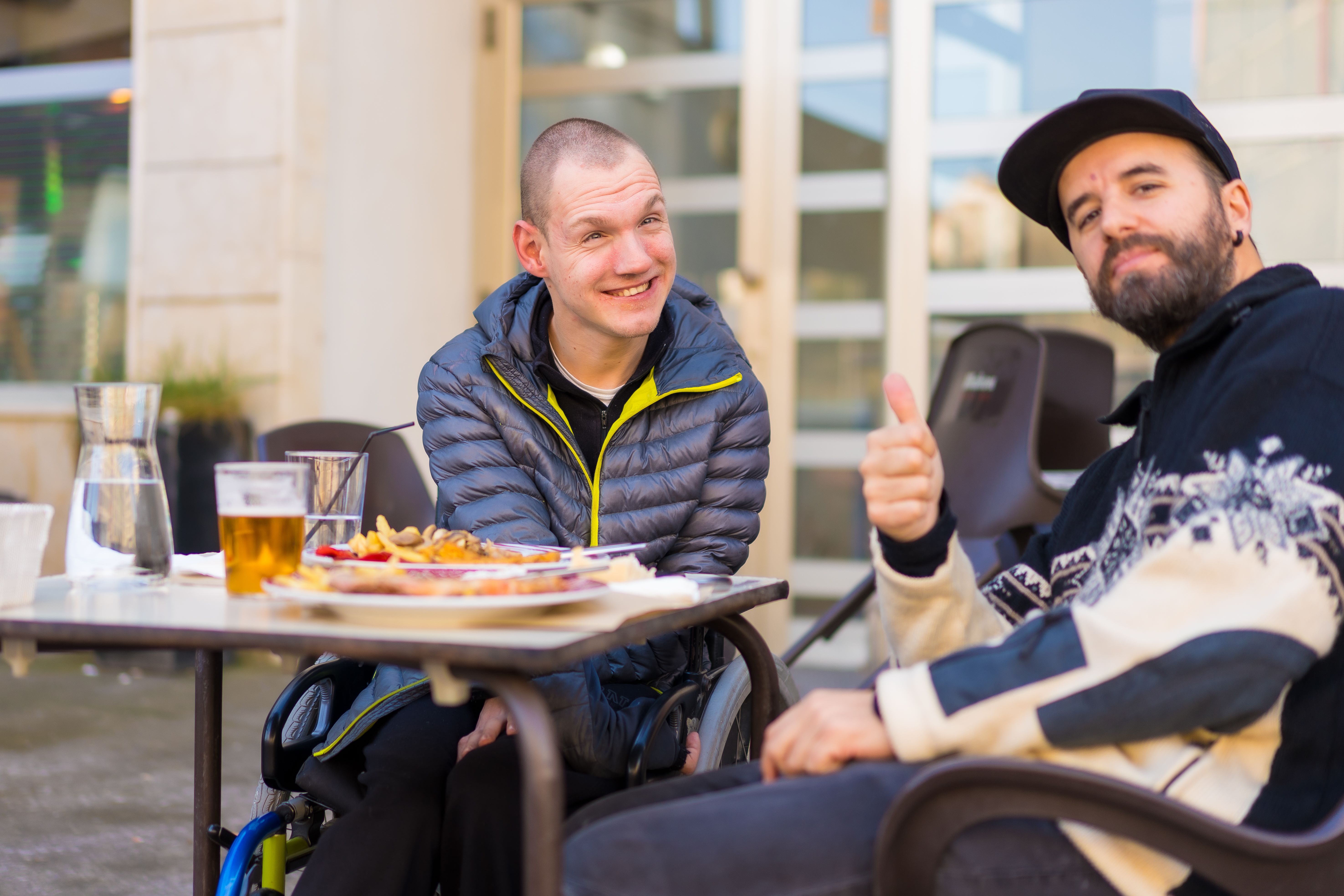 Cheerful young man in a wheelchair with his companion enjoying their meal outdoors on a sunny day